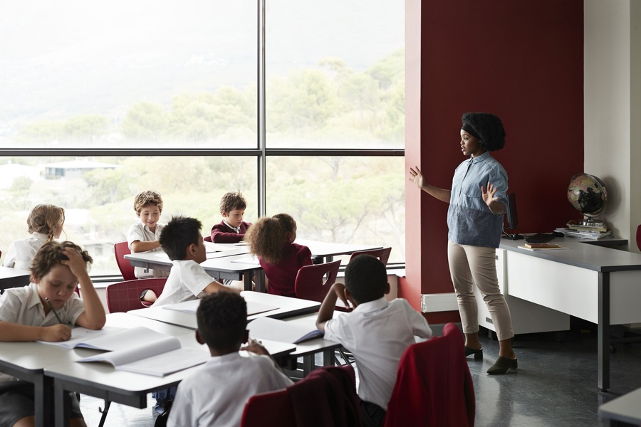 Professora de pé ensina alunos sentados em sala de aula moderna com janelas amplas.
