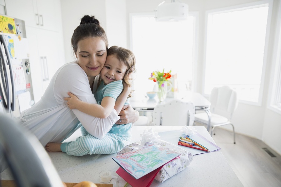 Mãe e filha se abraçando carinhosamente na cozinha, com desenhos e materiais de arte na bancada.