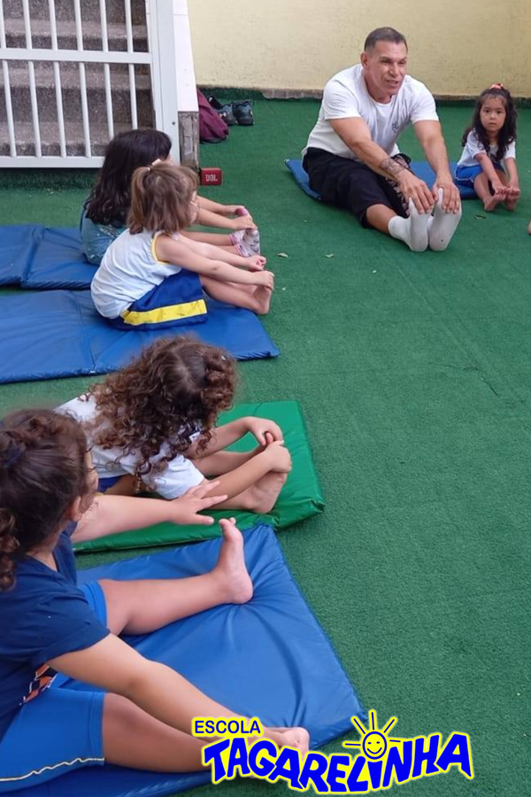Professor e crianças alongando em tapetes azuis e verdes durante aula de educação física escolar.