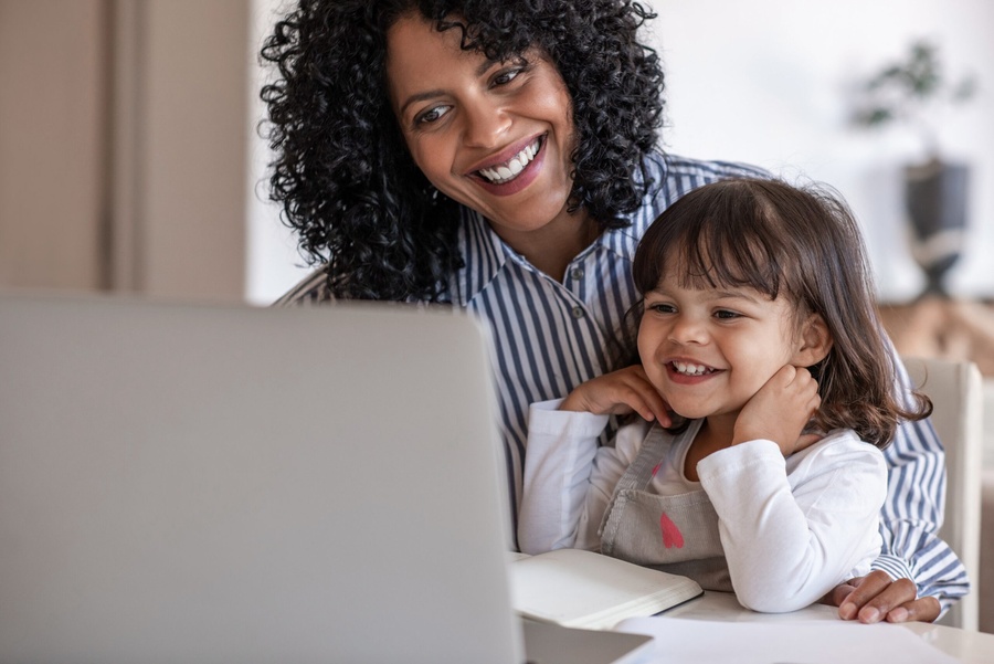 Mãe e filha sorrindo, olhando para um laptop branco em uma mesa.