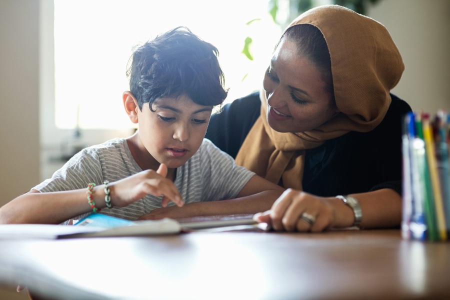Jovem garoto e mulher com hijab olhando um livro aberto em uma mesa de madeira clara.