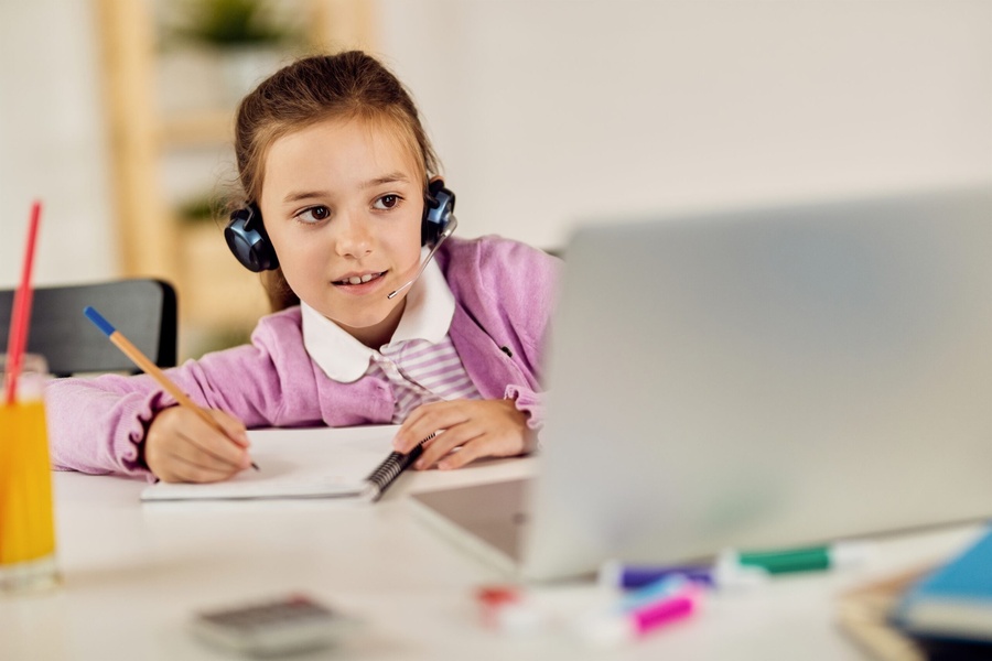 Menina com headset escrevendo em caderno, participando de aula online com laptop.