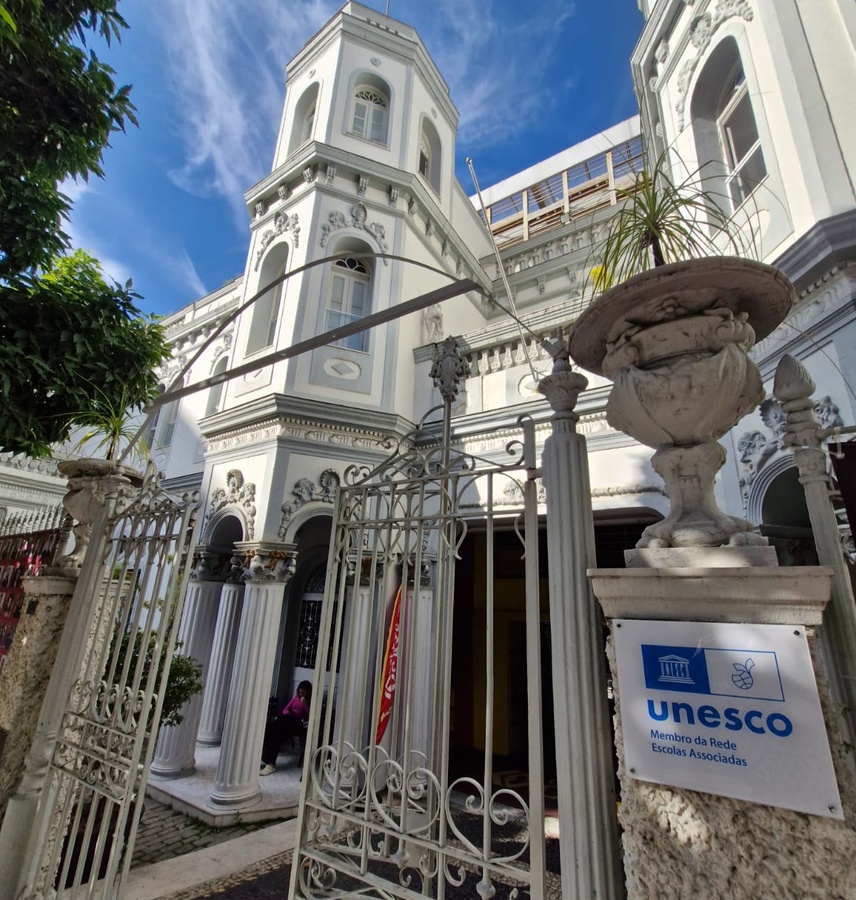 Edifício branco ornamentado com torre, portão metálico aberto e placa da UNESCO.