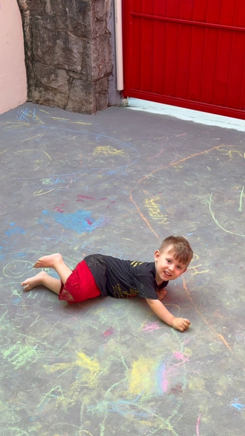 Menino sorridente de bruços em chão com desenhos coloridos de giz.