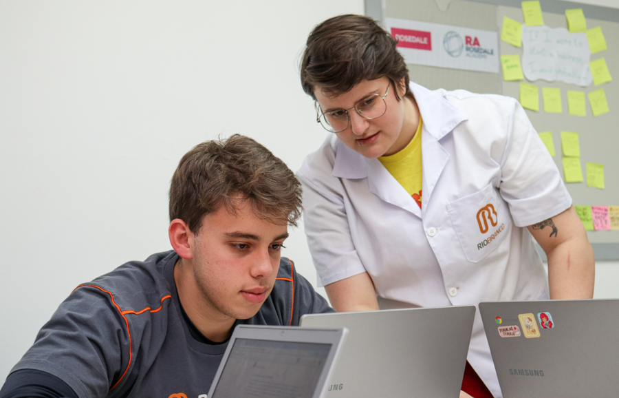 Duas pessoas colaboram em laptops, uma delas em jaleco branco, em ambiente educacional.
