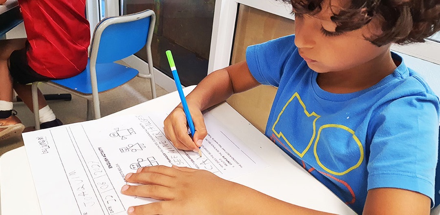 Criança com cabelo cacheado escrevendo em uma atividade escolar com lápis na mesa.