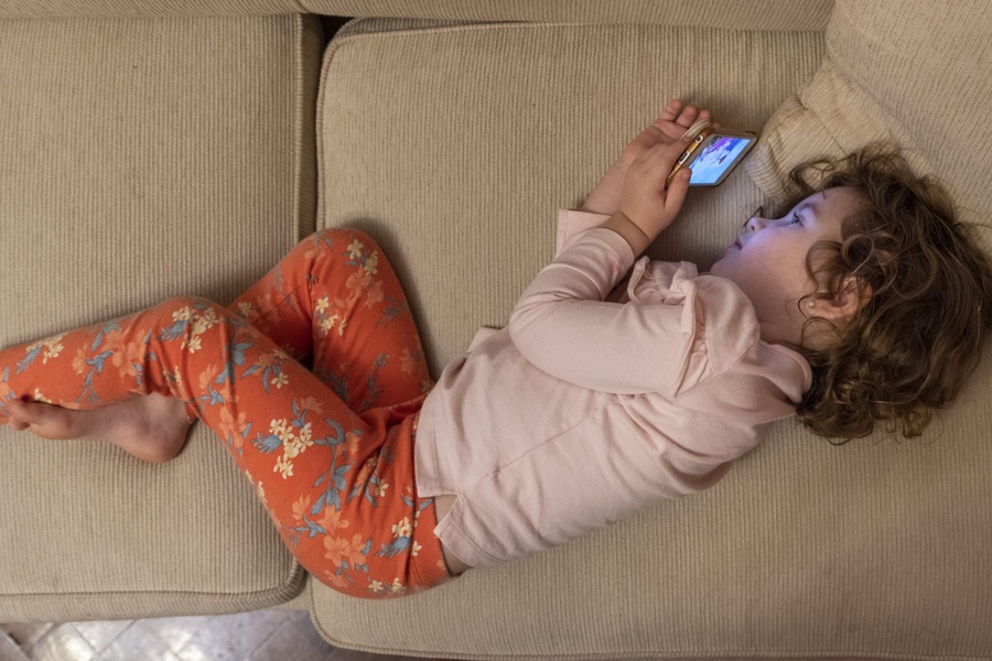 Criança deitada no sofá, usando um smartphone com a tela acesa, focada no aparelho.