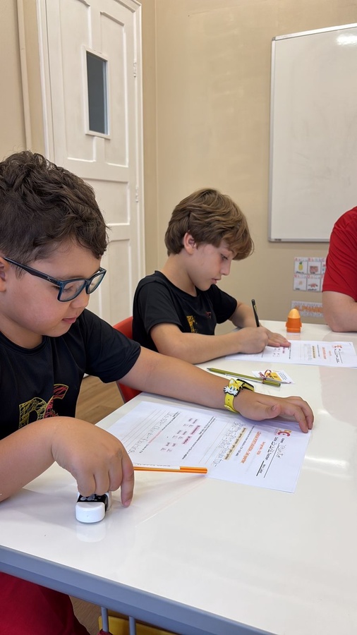 Dois meninos em uma mesa de aula focados em escrever e desenhar em papéis.