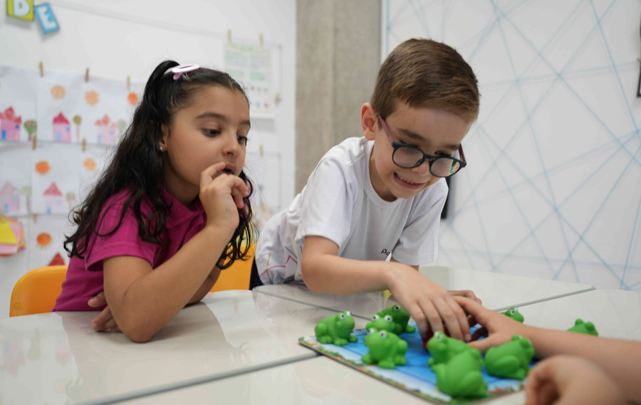 Duas crianças, um menino de óculos e uma menina, brincando com um jogo de sapos verdes em uma mesa.