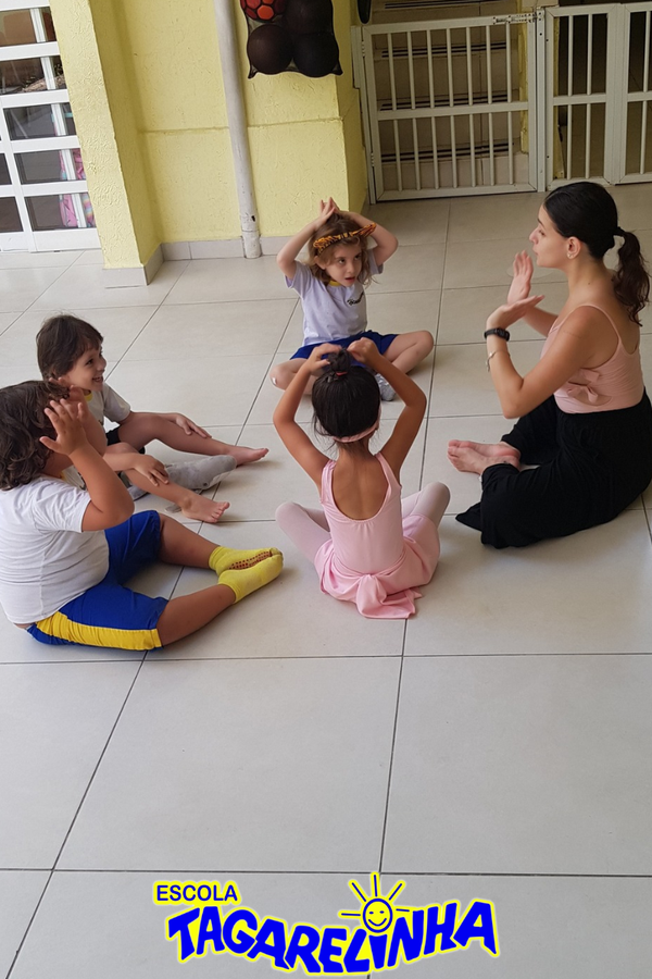 Crianças e professora sentadas no chão fazendo gestos com as mãos em atividade lúdica.