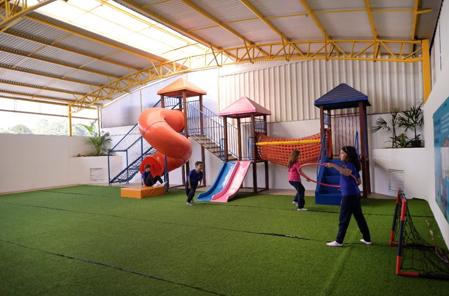 Crianças brincando em um playground coberto com grama sintética, escorregadores e estruturas de escalada.