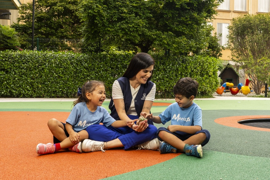 Educadora e duas crianças felizes em um playground, segurando uma pequena planta com terra.