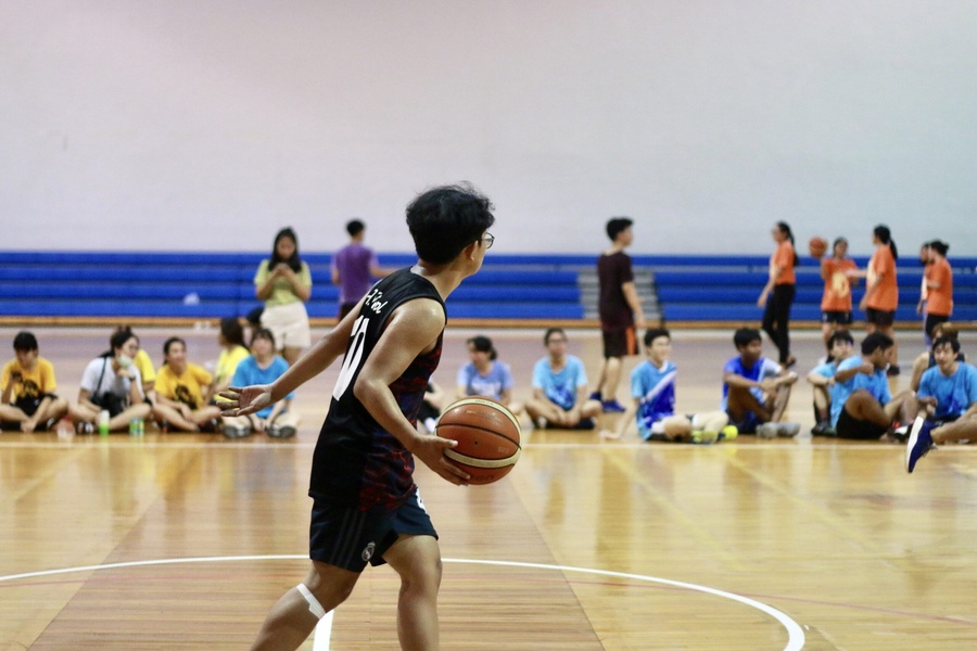 Jovem driblando bola de basquete em quadra de madeira com pessoas sentadas ao fundo.