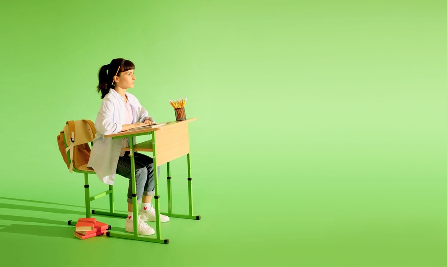 Estudante sentada em carteira escolar verde, com lápis, livros e mochila, olhando para o lado em fundo vibrante.