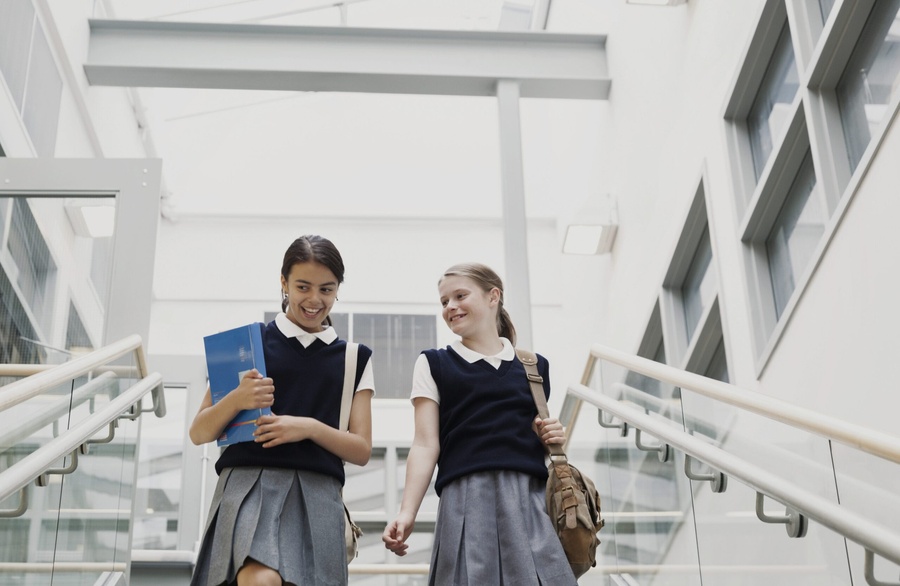 Duas estudantes uniformizadas descendo escadas modernas, sorrindo e carregando materiais escolares.