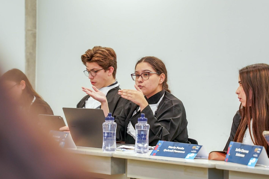 Jovens estudantes em becas acadêmicas participando de um debate ou apresentação formal.