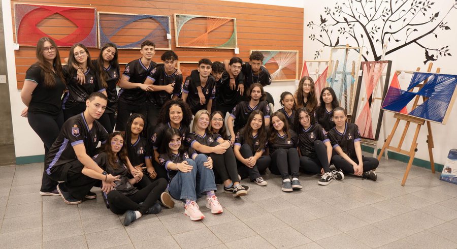 Grupo de jovens estudantes em camisas pretas posando em exposição de arte de fios coloridos.
