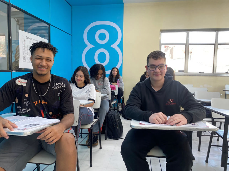 Estudantes sorrindo em sala de aula com carteiras e lousa azul decorada.