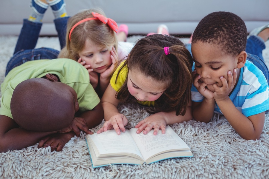 Crianças diversas deitadas em um tapete fofo, lendo atentas um livro.