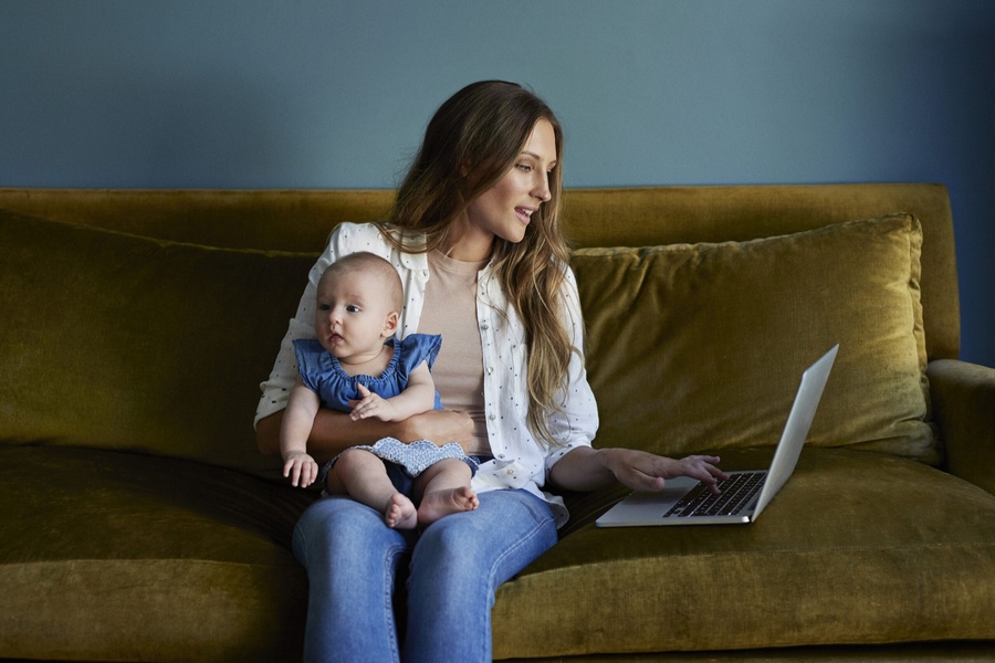 Mãe segura bebê enquanto usa laptop no sofá verde-oliva com parede azul.