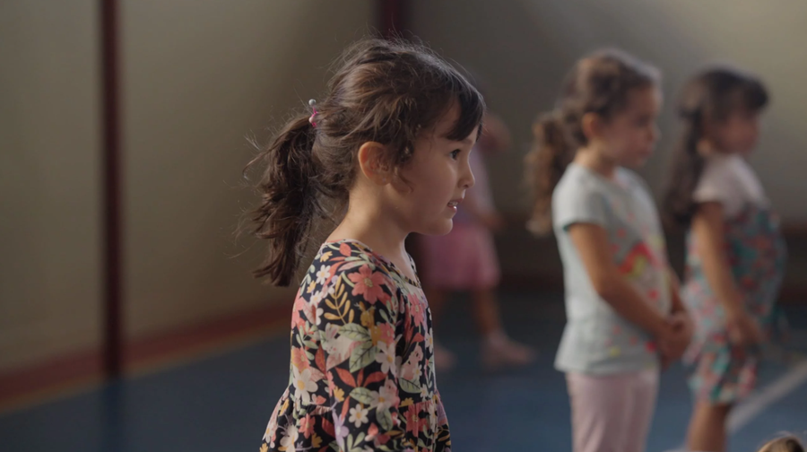 Menina com cabelo preso e vestido floral observando, com outras crianças desfocadas ao fundo.