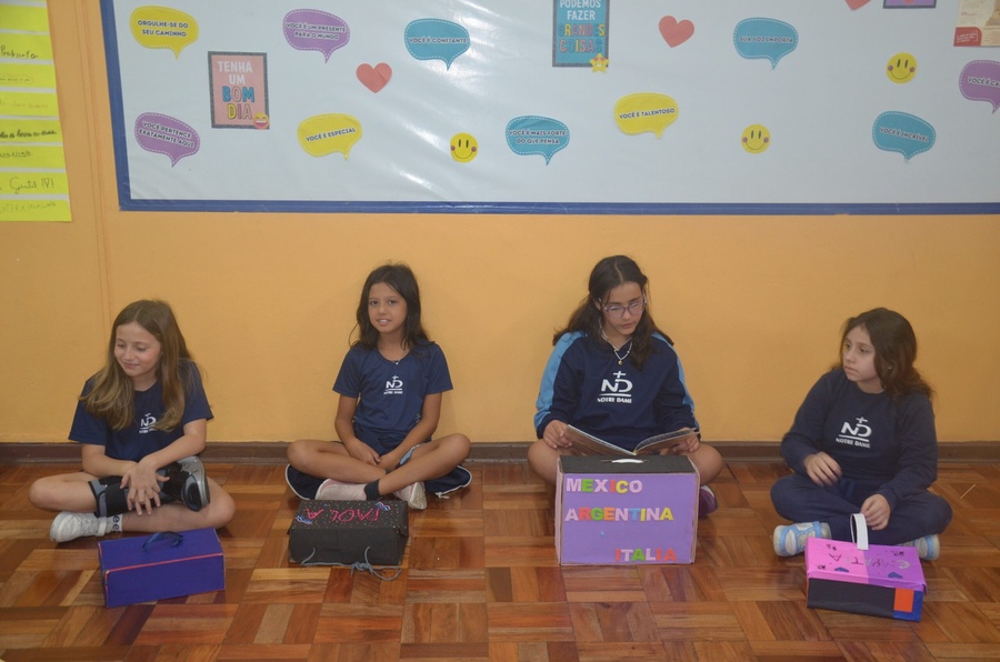 Quatro alunas sentadas no chão da sala de aula com caixas e lendo um livro.