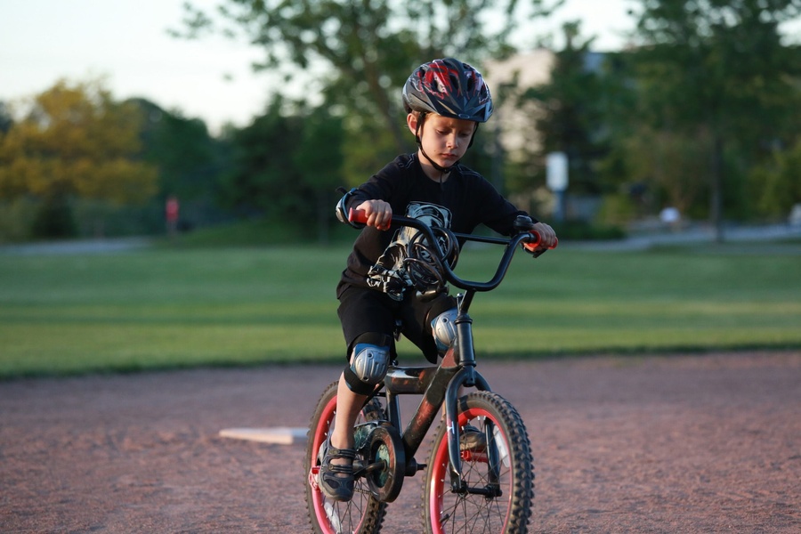 Menino usando capacete e joelheiras em bicicleta preta e vermelha em campo de terra.