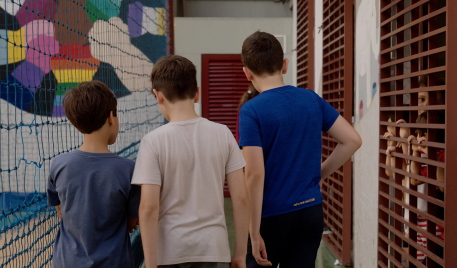 Três meninos de costas caminham; uma criança observa através de grades ao lado.