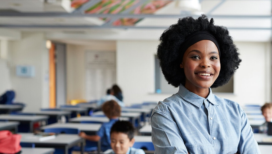 Mulher negra sorridente com cabelo afro em sala de aula, braços cruzados.