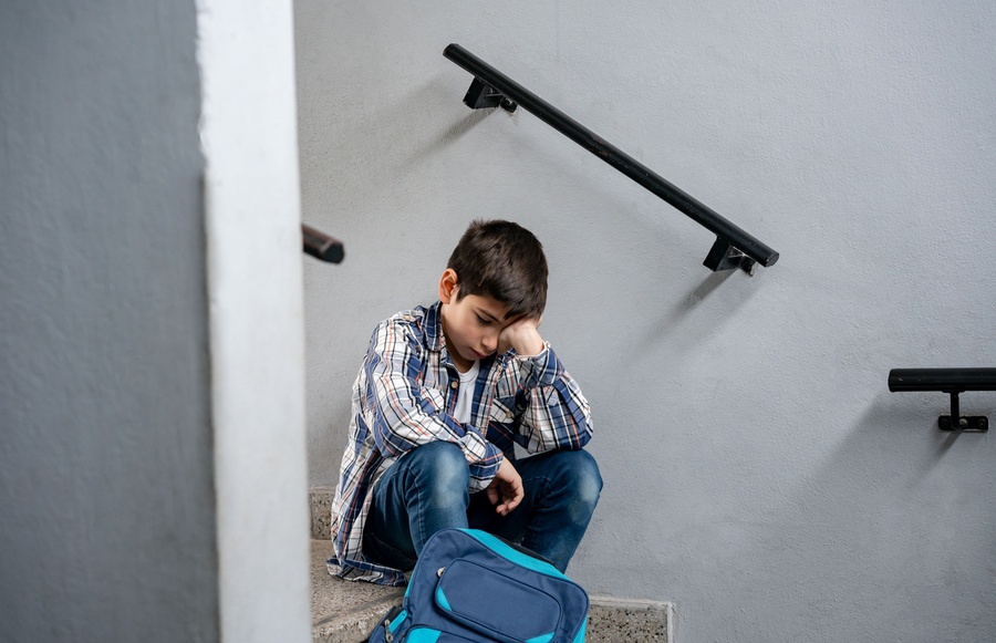 Menino com expressão triste sentado em escada, com mochila ao lado e cabeça na mão.