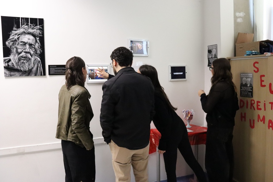 Pessoas observam exposição interativa sobre idosos, direitos humanos, com fotos e telas digitais.